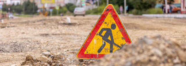 Afbeelding van een driehoekig verkeersbord met een rode rand en een mannetje dat aan het werk is in het midden. Het bord ligt op een opengebroken straat.